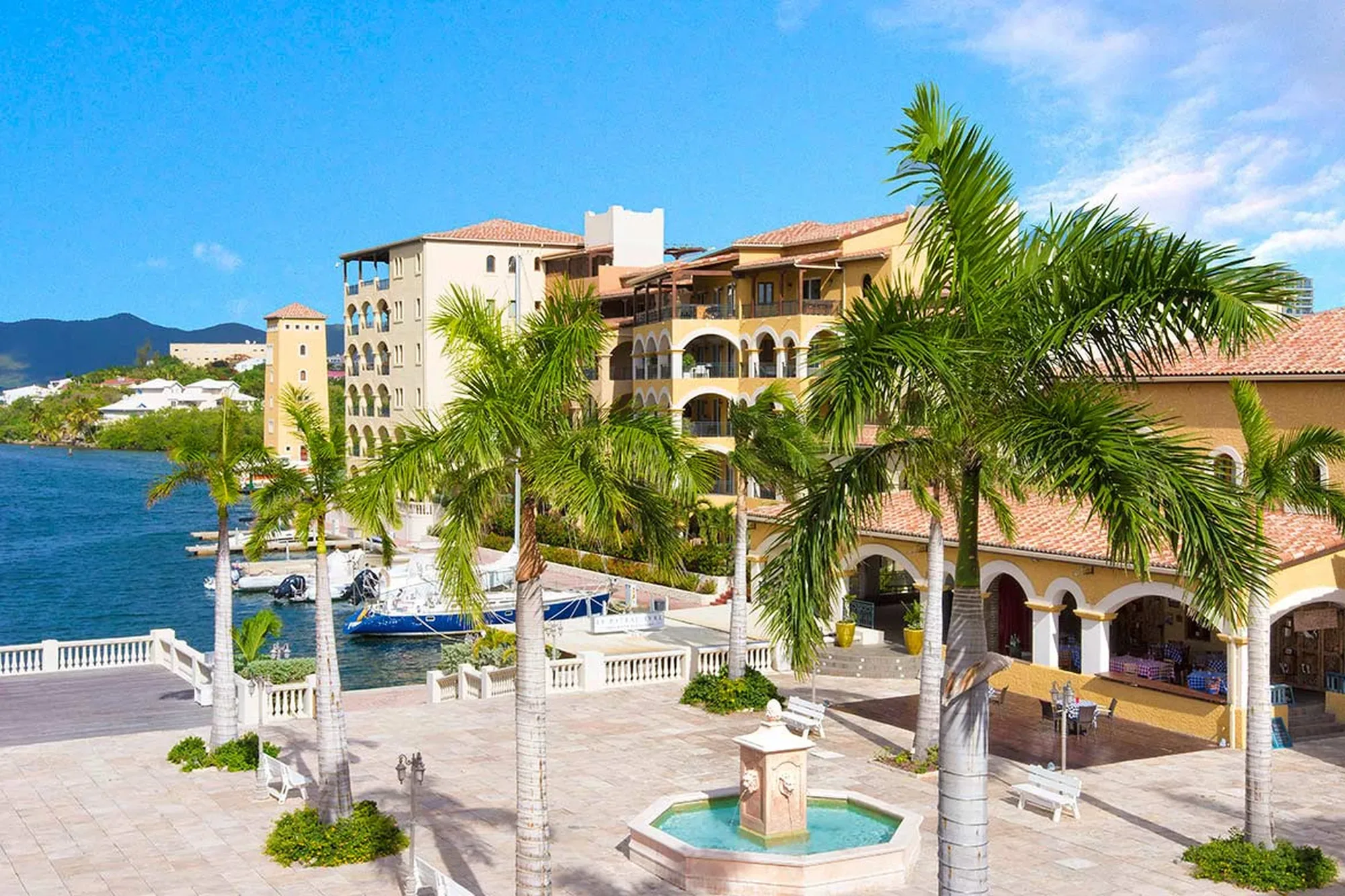 A grand waterfront plaza anchored by an ornate fountain — where Mediterranean colonnades meet private yacht moorings and sun-drenched Caribbean waters.