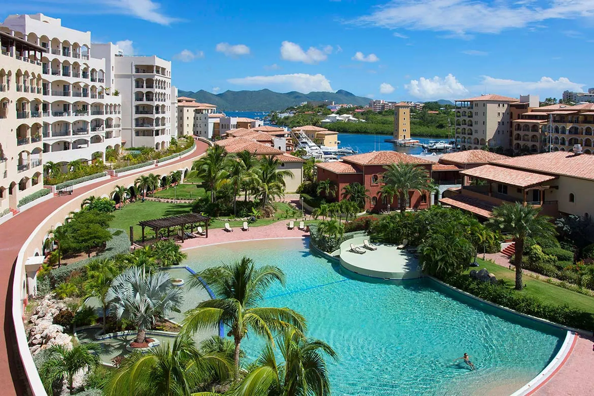 A crystalline lagoon-style pool unfurls beneath the Caribbean sky, framed by Mediterranean architecture and lush tropical landscaping that whispers of private paradise.