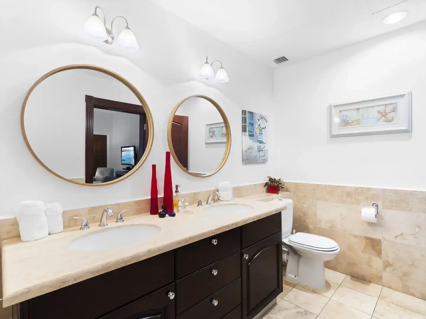 Bright master bathroom with a dark wood double vanity, two mirrors, and Mediterranean courtyard artwork on the walls.