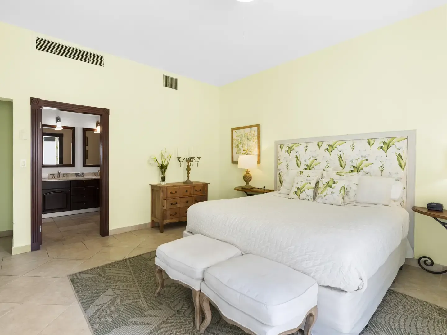 Light-filled bedroom with a botanical print headboard in greens and yellows, white benches, and yellow floral accents on the dresser.