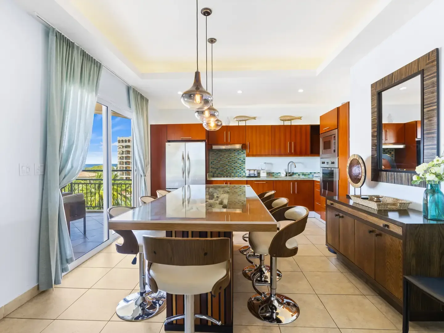 Bright open kitchen with warm wood cabinetry, a glass-top island with seashell display, and stunning sea views through sliding glass doors.
