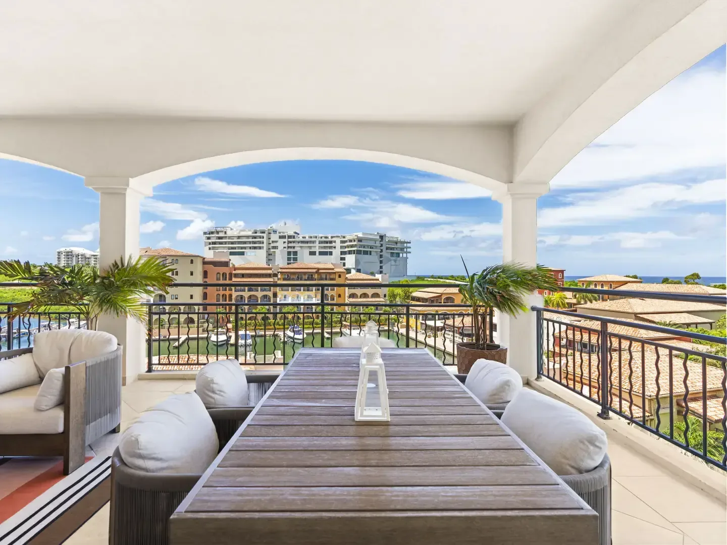 Sophisticated covered terrace with a grey weathered-wood dining table and plush lounge area, flanked by tall palm trees and a marina backdrop.