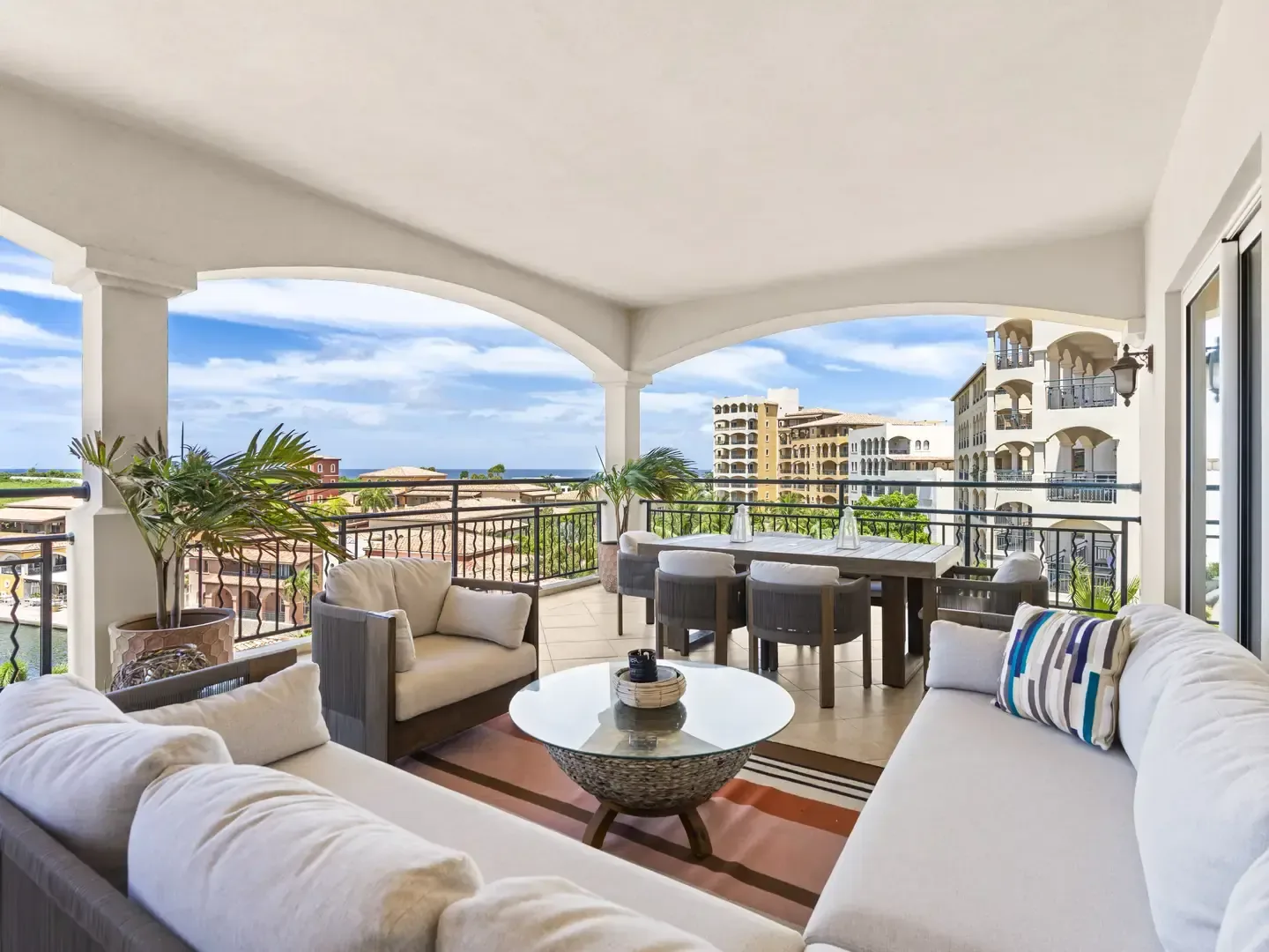 Spacious covered terrace with a plush L-shaped sectional, lounge chairs, and a dining area framed by elegant white arches and tropical palms.