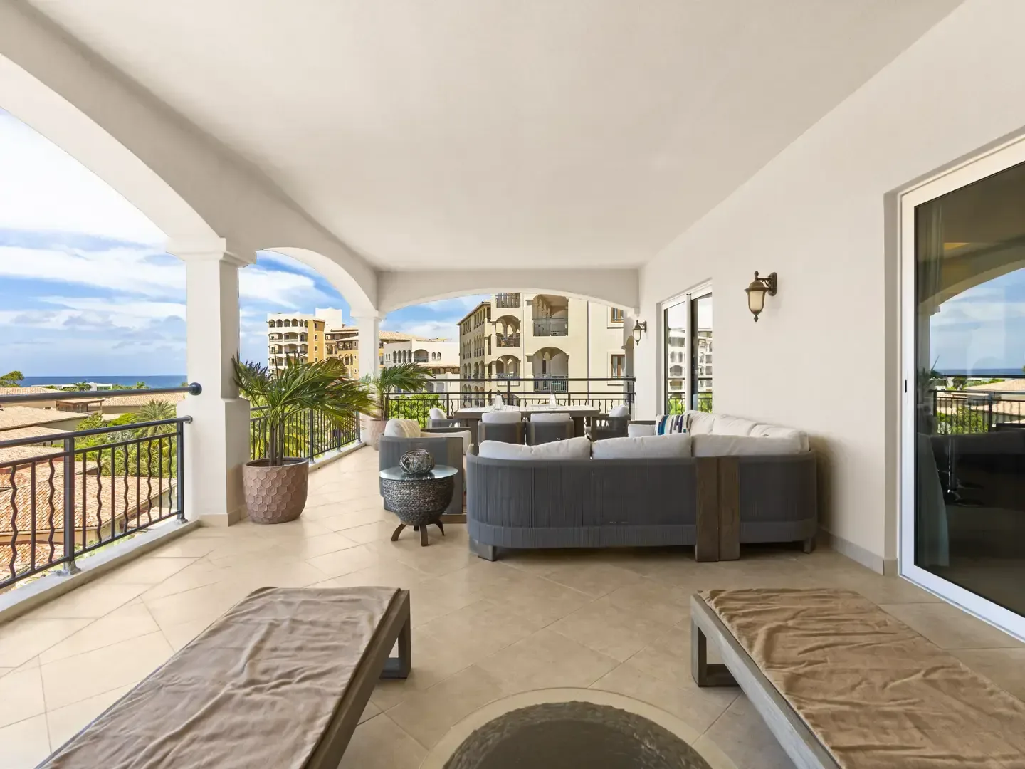 Airy covered terrace with lounge chairs and an outdoor dining set, accented by potted palms and framed by arched openings with ocean views.