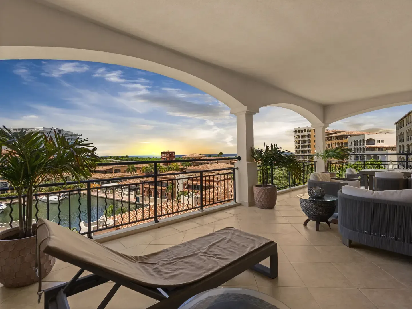 Grand covered balcony at sunset with curved sofas, a lounge chair, and a dining set overlooking a turquoise marina and the open ocean.
