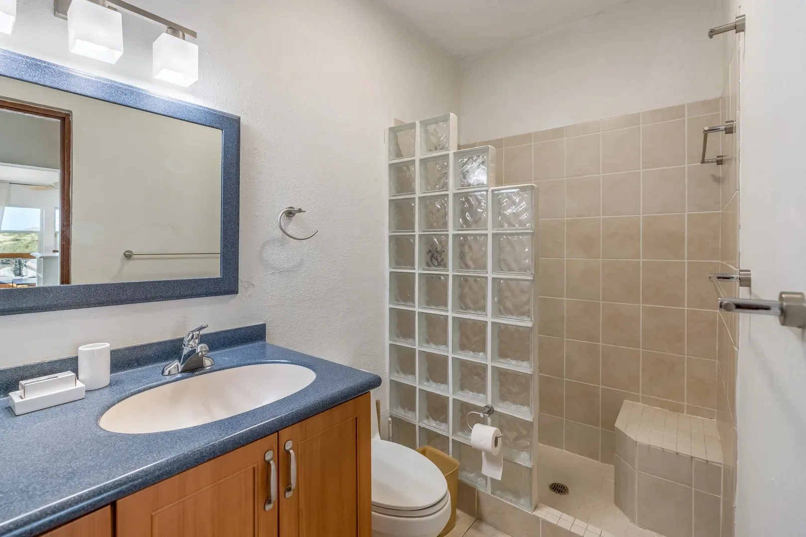 Bathroom with blue sink countertop, wooden vanity, glass block shower wall, beige tile shower, and mirror reflecting outdoor view