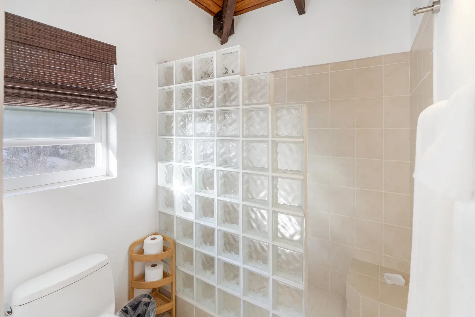 Bathroom with glass block wall, white toilet, wooden shelf, beige tile shower, and window with bamboo blind