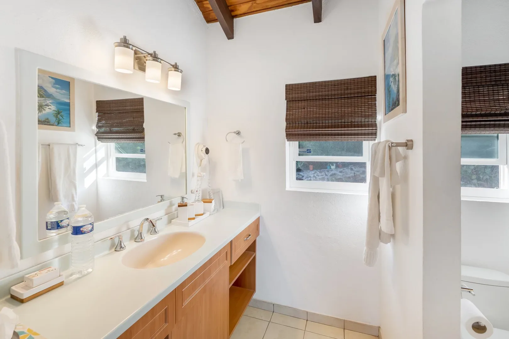 Bright bathroom with white countertop, light wood vanity, large mirror, bamboo window blinds, and tropical landscape artwork on the wall