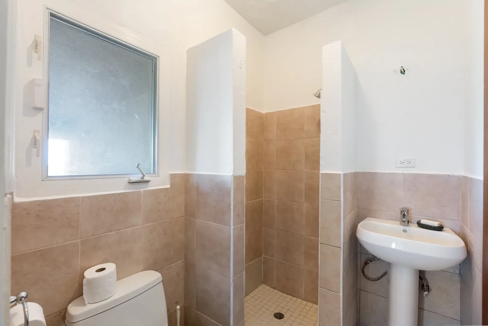 Bright bathroom with beige tile walls, white sink, toilet, shower area, and frosted window