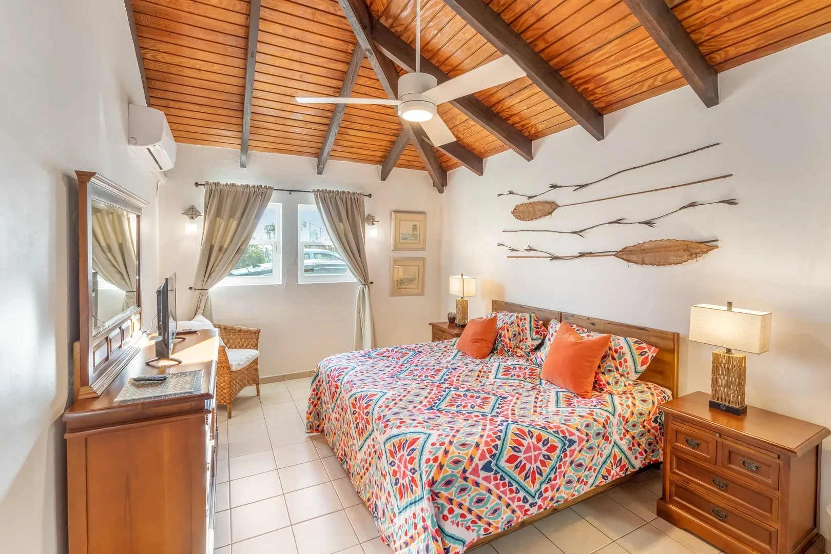 Bright bedroom with large bed, colorful geometric bedspread, wooden nightstands, wicker chair, two windows with beige curtains, and exposed wooden ceiling beams