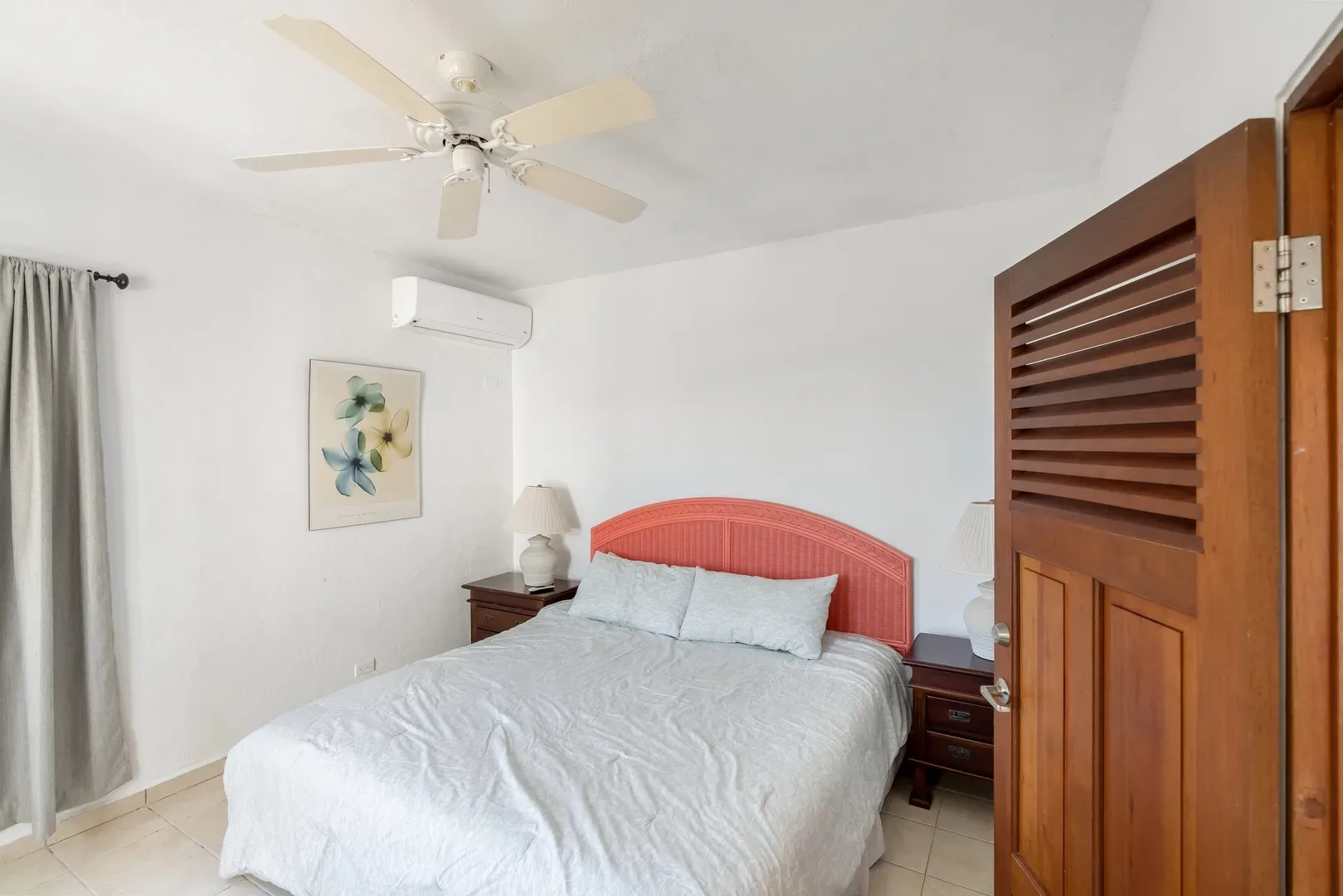 Bright bedroom with coral headboard, white linens, wooden nightstands, ceiling fan, floral artwork, air conditioning, and louvered wooden door