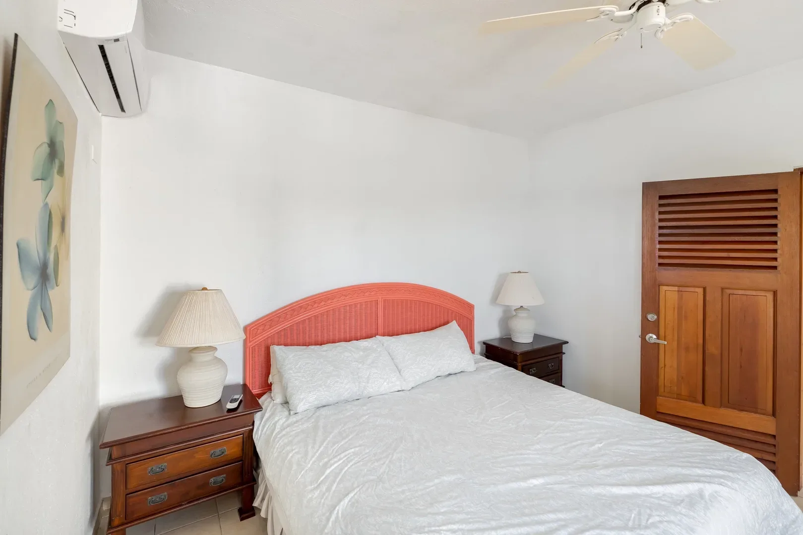St. Maarten villa bedroom with coral woven headboard, white linens, wooden nightstands, ceiling fan, and floral artwork