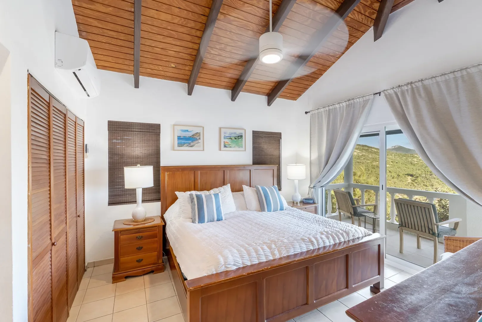 Spacious bedroom with wooden bed frame, white bedding, blue-striped pillows, nightstands with lamps, ceiling fan, and sliding glass doors leading to a balcony with chairs and a view of green hills