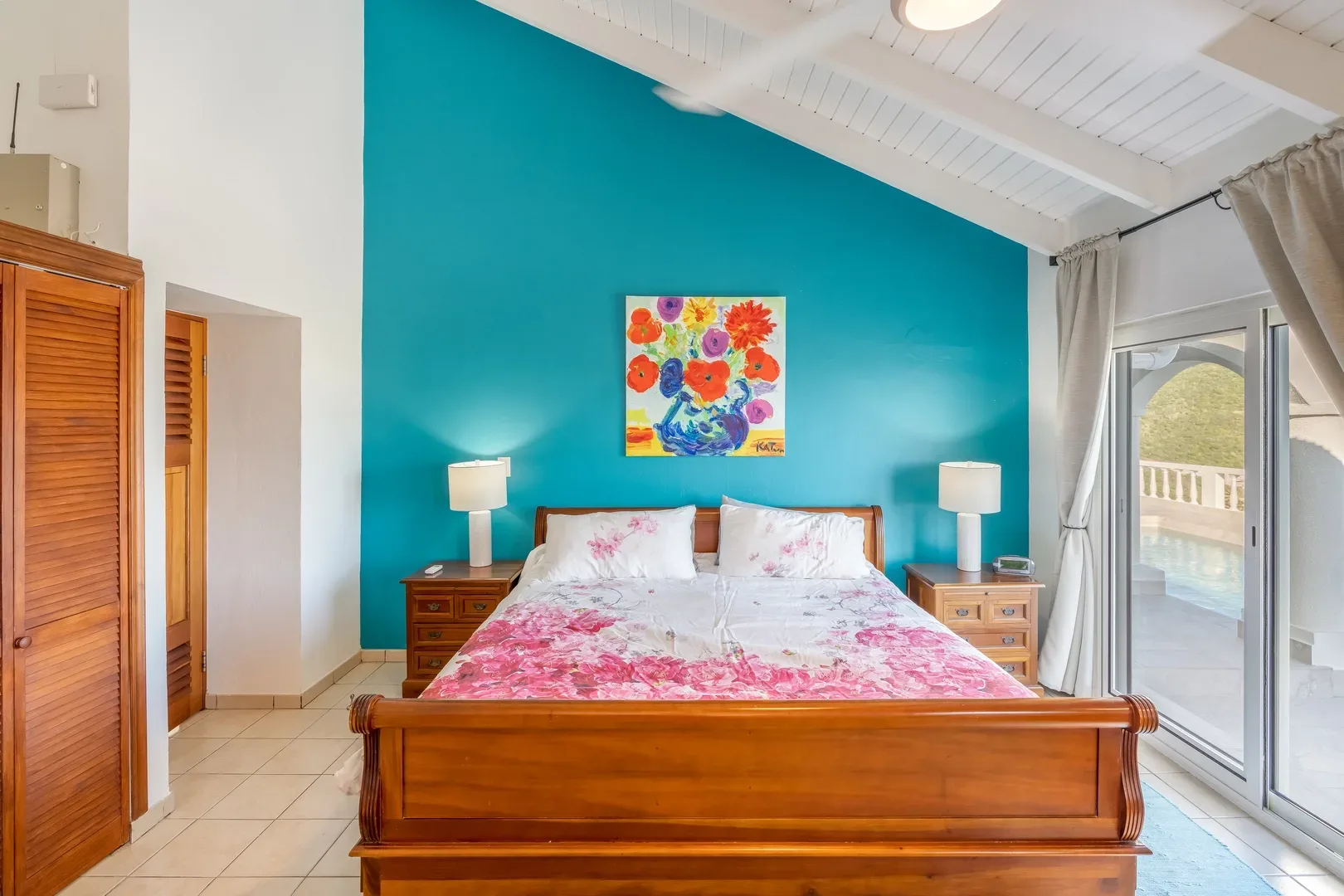 Bright master bedroom with wooden sleigh bed, pink floral bedding, turquoise accent wall, and sliding glass doors overlooking a pool and tropical landscape
