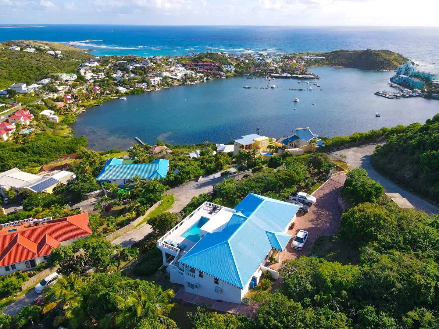 Aerial view of a white villa with a bright blue roof and pool, overlooking a turquoise bay with boats, surrounded by lush greenery and coastal homes in St. Maarten