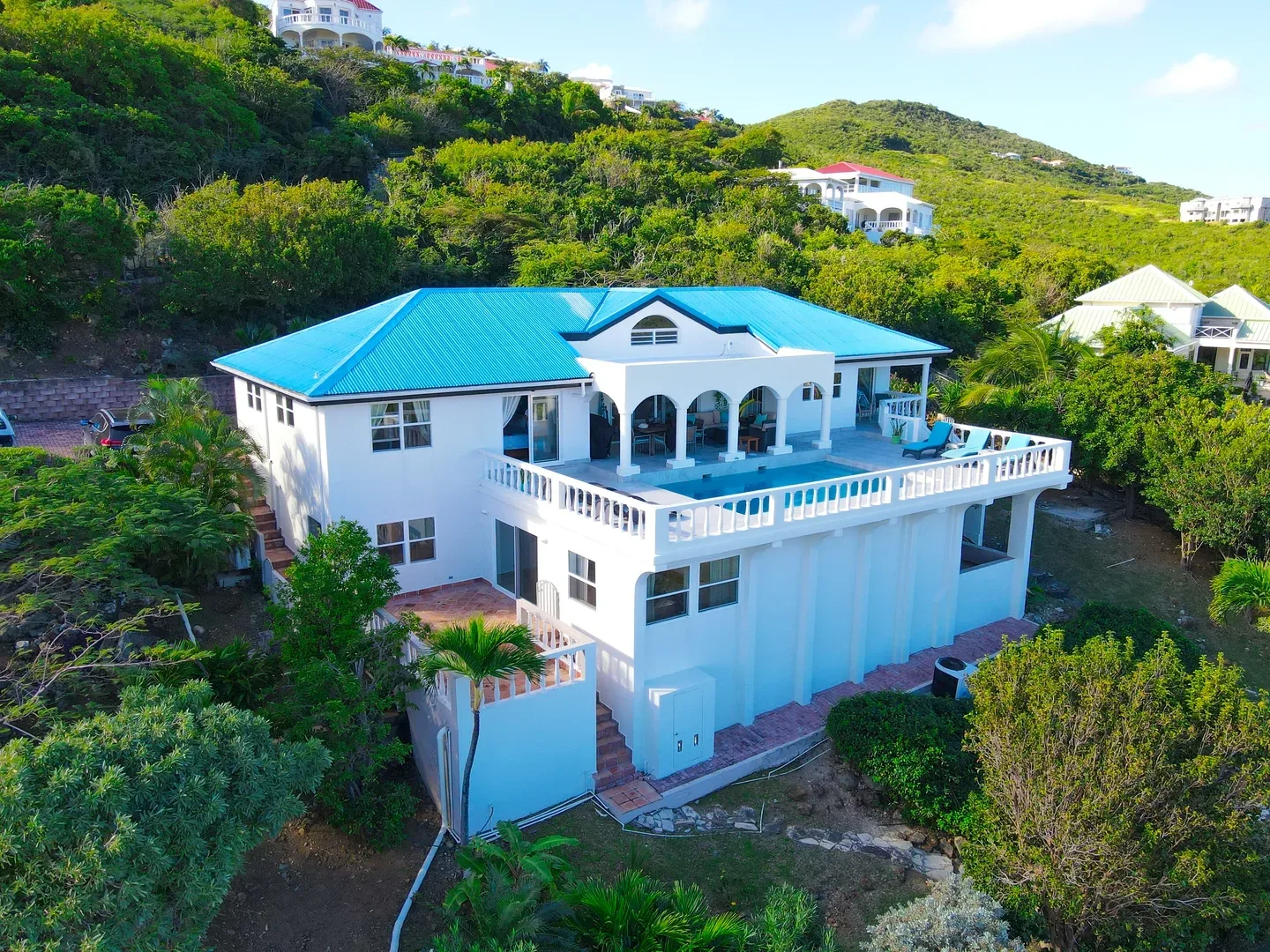 Aerial view of a white luxury villa with a bright blue roof and upper-level swimming pool, surrounded by lush green hills in St. Maarten