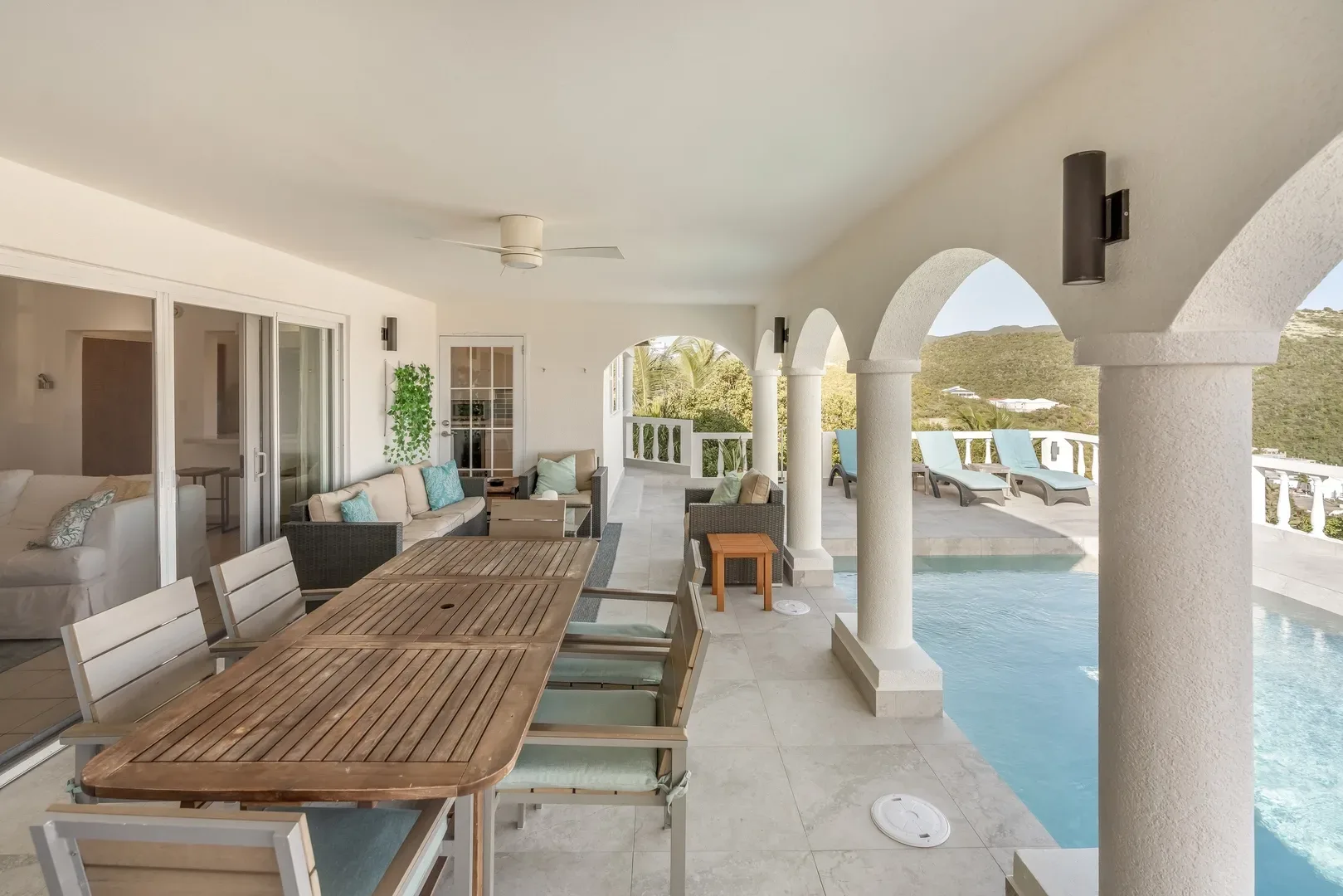 Covered outdoor terrace at a St. Maarten villa with wooden dining table, blue-cushioned chairs, wicker seating, white arches, swimming pool, and green hill views
