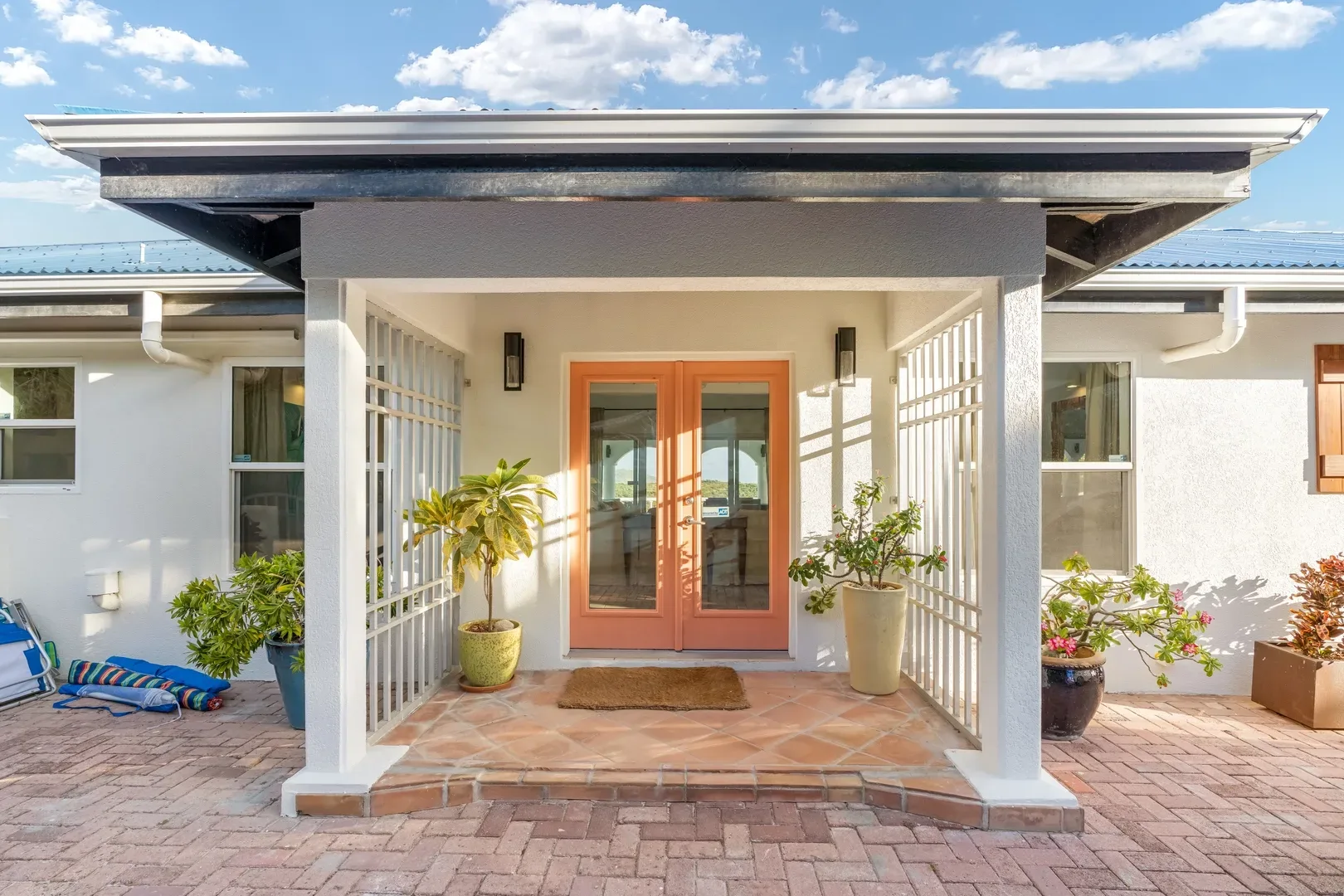 Front entrance of St. Maarten villa with orange double glass doors, covered porch, potted plants, brick patio, and blue sky