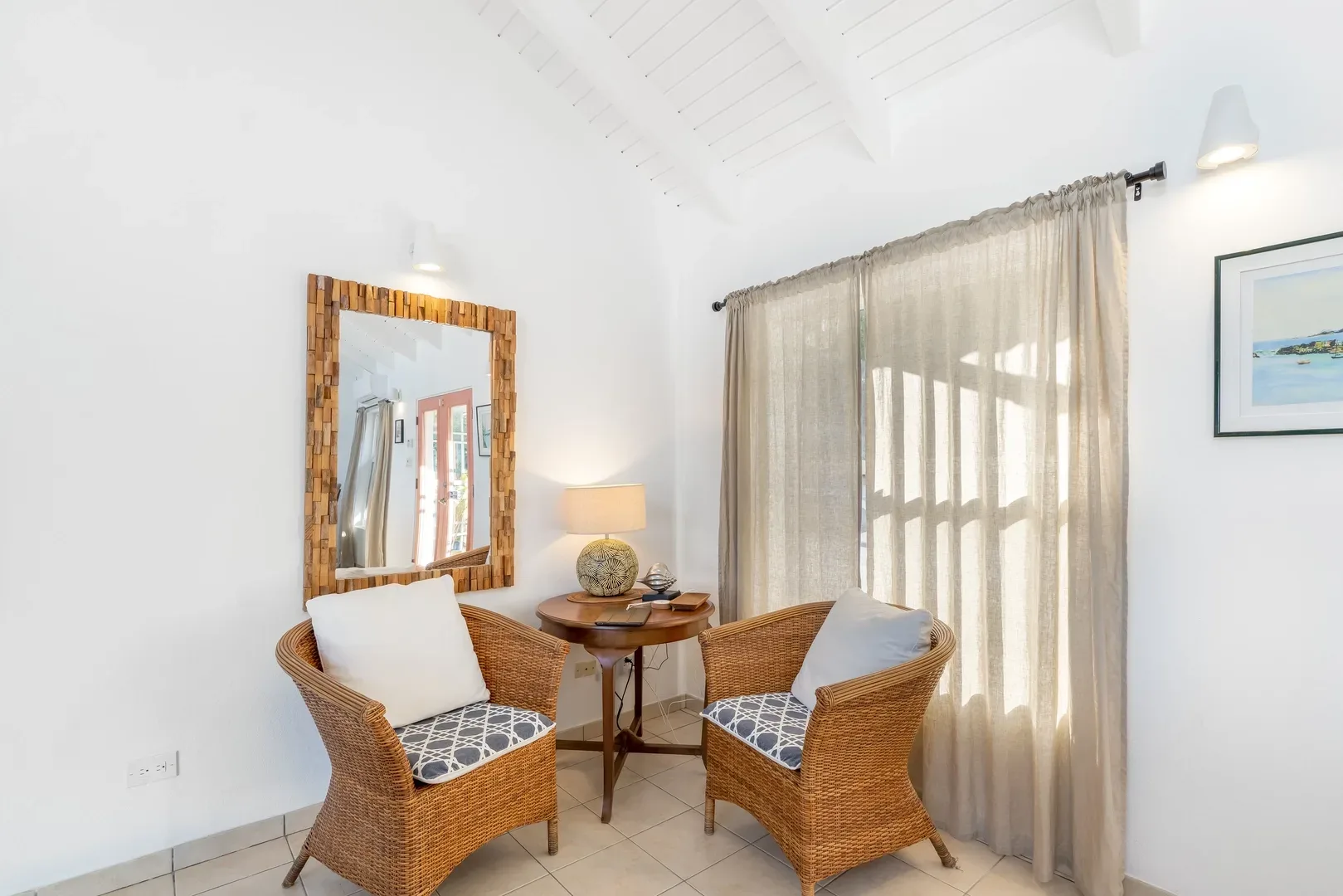 Cozy sitting area with two wicker chairs, patterned cushions, wooden table, mirror, and beige curtains in a bright villa room