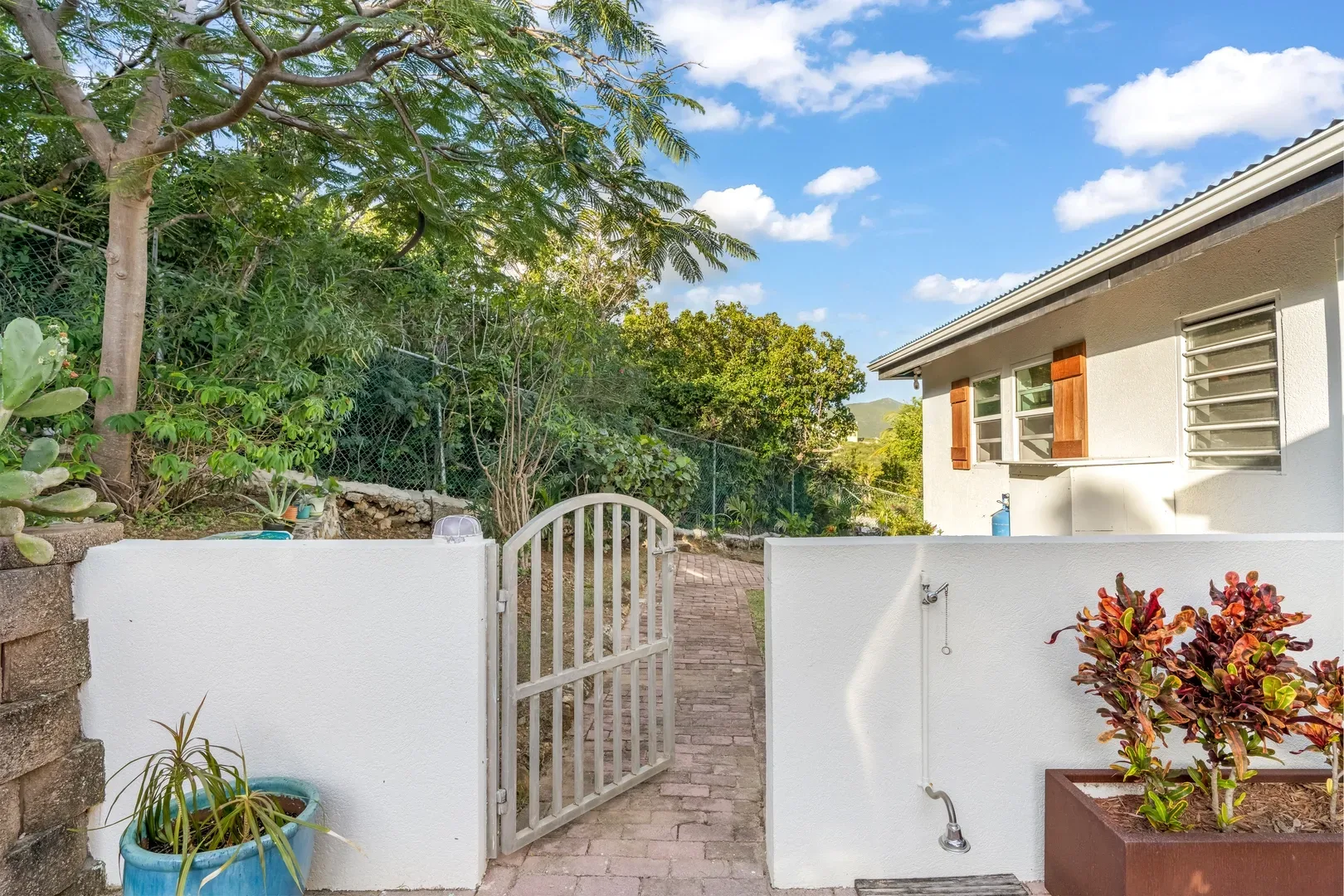 White villa exterior in St. Maarten with open metal gate, brick pathway, outdoor shower, tropical plants, and blue sky with clouds