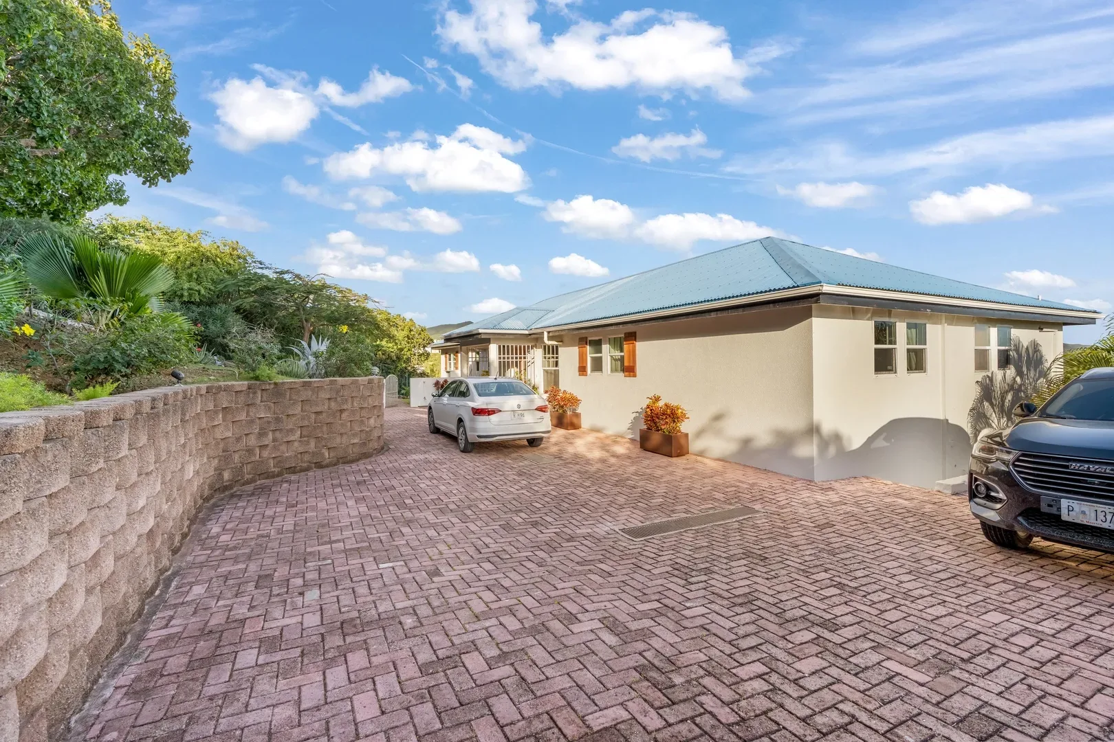 Exterior of a beige villa with blue roof in St. Maarten, featuring brick driveway, two parked cars, tropical landscaping, and bright blue sky with clouds