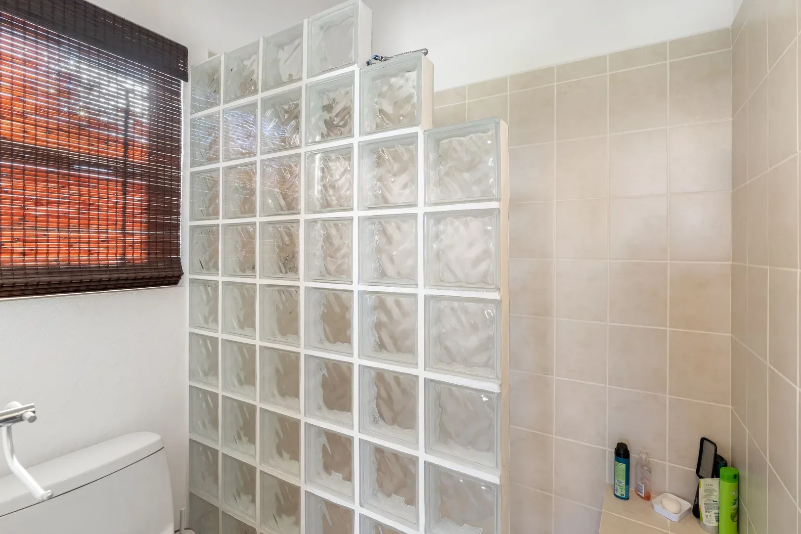 Bathroom with glass block partition, beige tile walls, white toilet, bamboo window blinds, and toiletries on counter