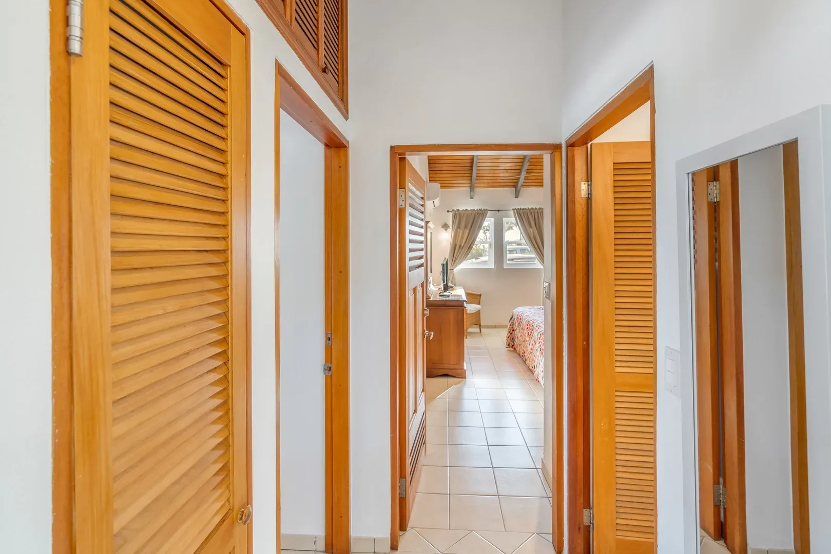 Hallway with wooden louvered doors leading to a bedroom with a bed, desk, and windows in a villa