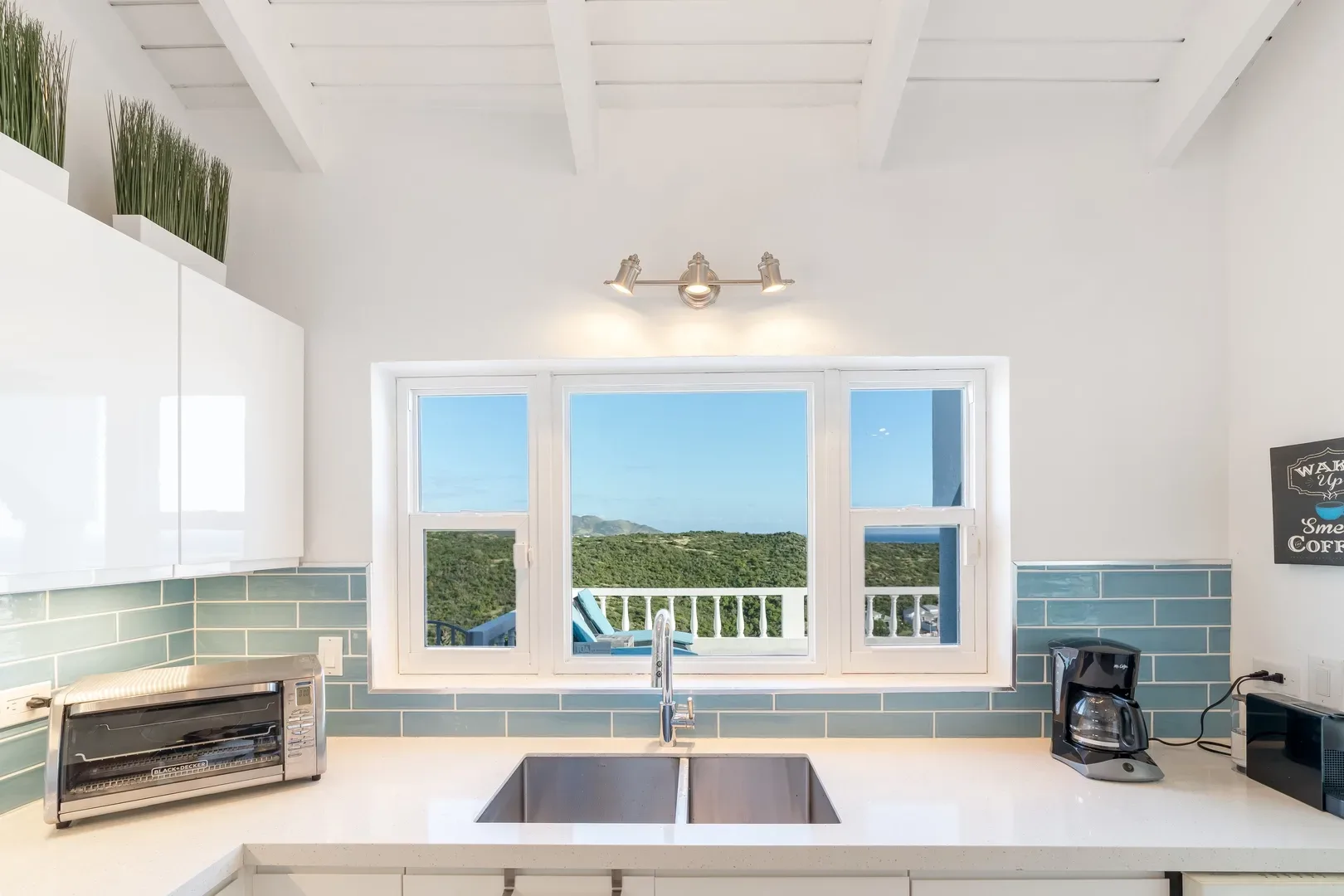 Bright modern kitchen with white cabinets, light blue tile backsplash, stainless steel sink, and large window showcasing ocean and hill views in St. Maarten villa