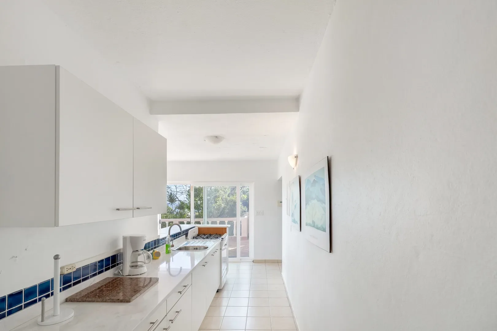 Spacious white kitchen with blue tile backsplash, white cabinets, and sliding glass doors leading to a balcony with ocean and tree views