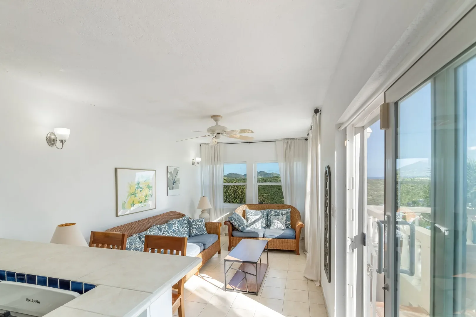 Bright open-concept living area with wicker sofas, blue patterned cushions, kitchen counter, and sliding glass doors leading to a balcony with mountain and greenery views