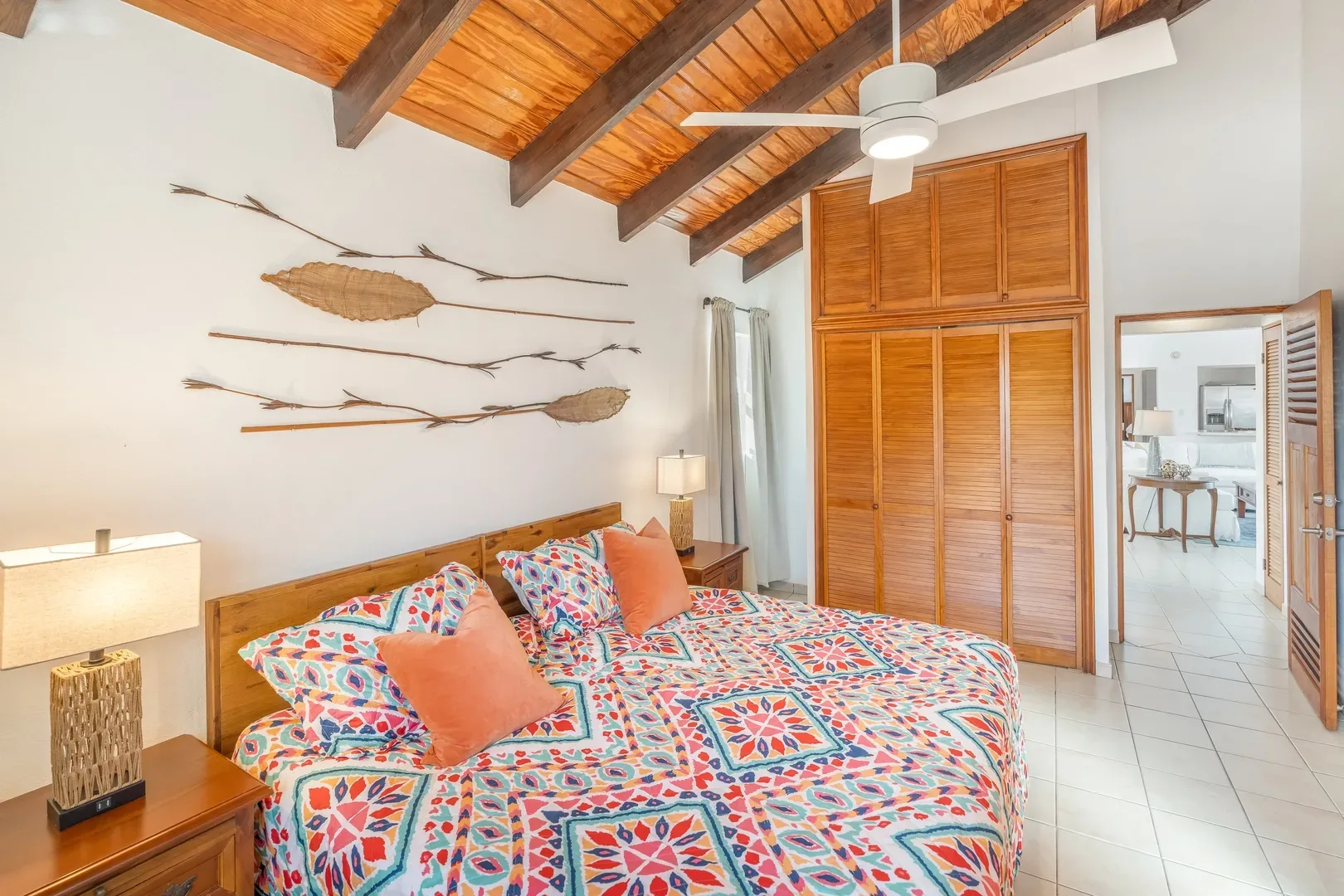 Bright master bedroom with colorful geometric bedspread, wooden ceiling beams, nightstands with lamps, and view of adjacent living area