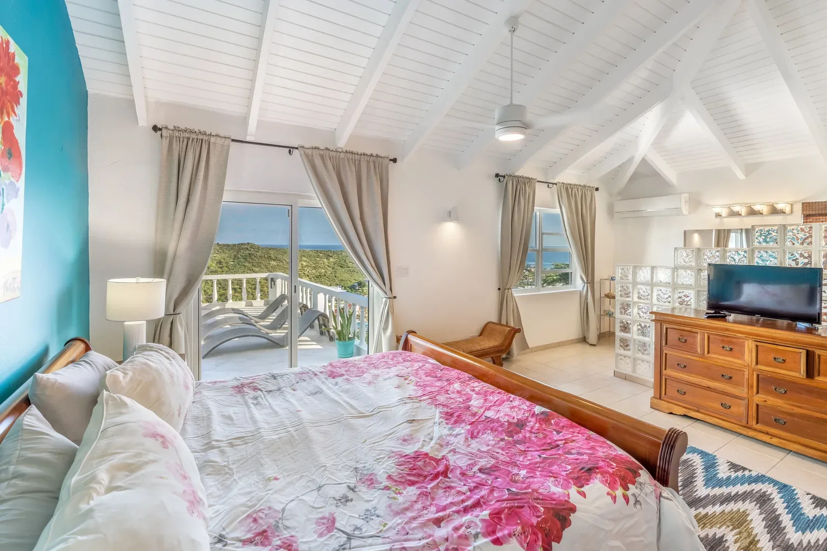 A spacious bedroom with a floral bed, turquoise accent wall, and sliding doors leading to a balcony with ocean and hill views