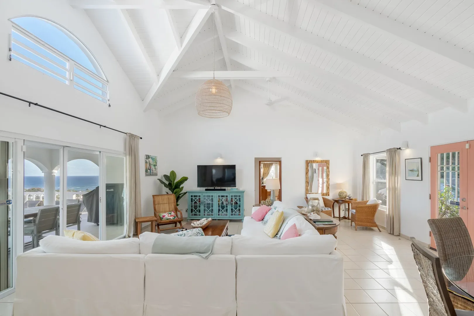 Spacious living room with white sectional sofa, turquoise cabinet, ocean view through glass doors, high vaulted ceiling, and natural light
