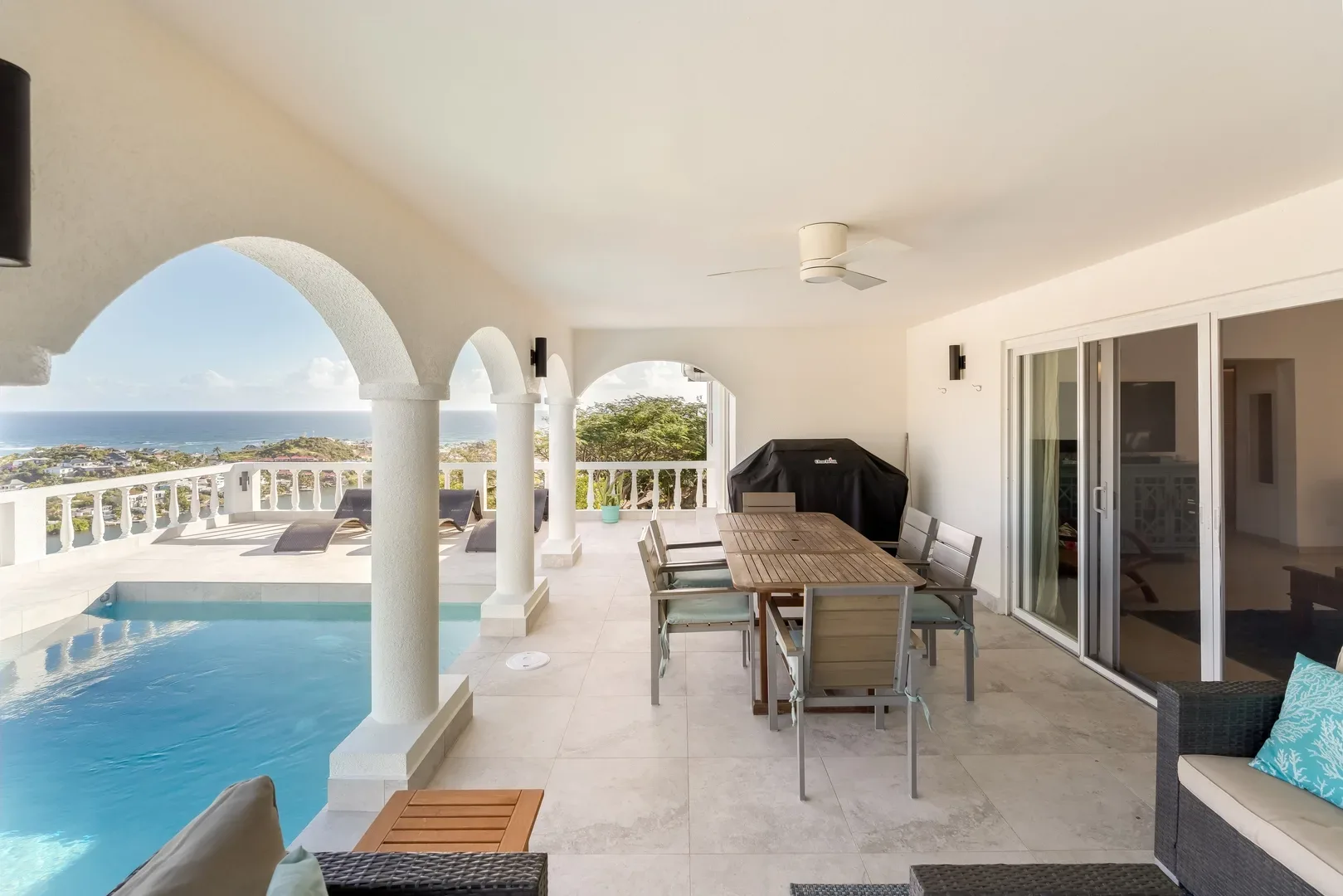Covered patio of a St. Maarten luxury villa with ocean view, pool, wooden dining table, and turquoise-cushioned seating