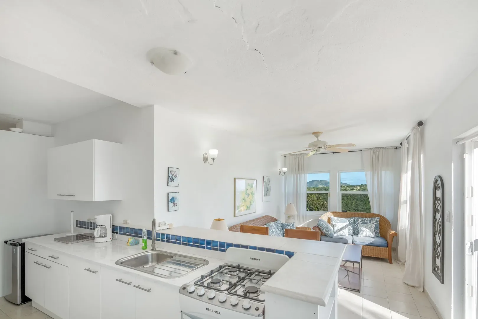 Open-concept kitchen and living area with white cabinets, wicker sofa, and mountain view through large windows in a St. Maarten villa
