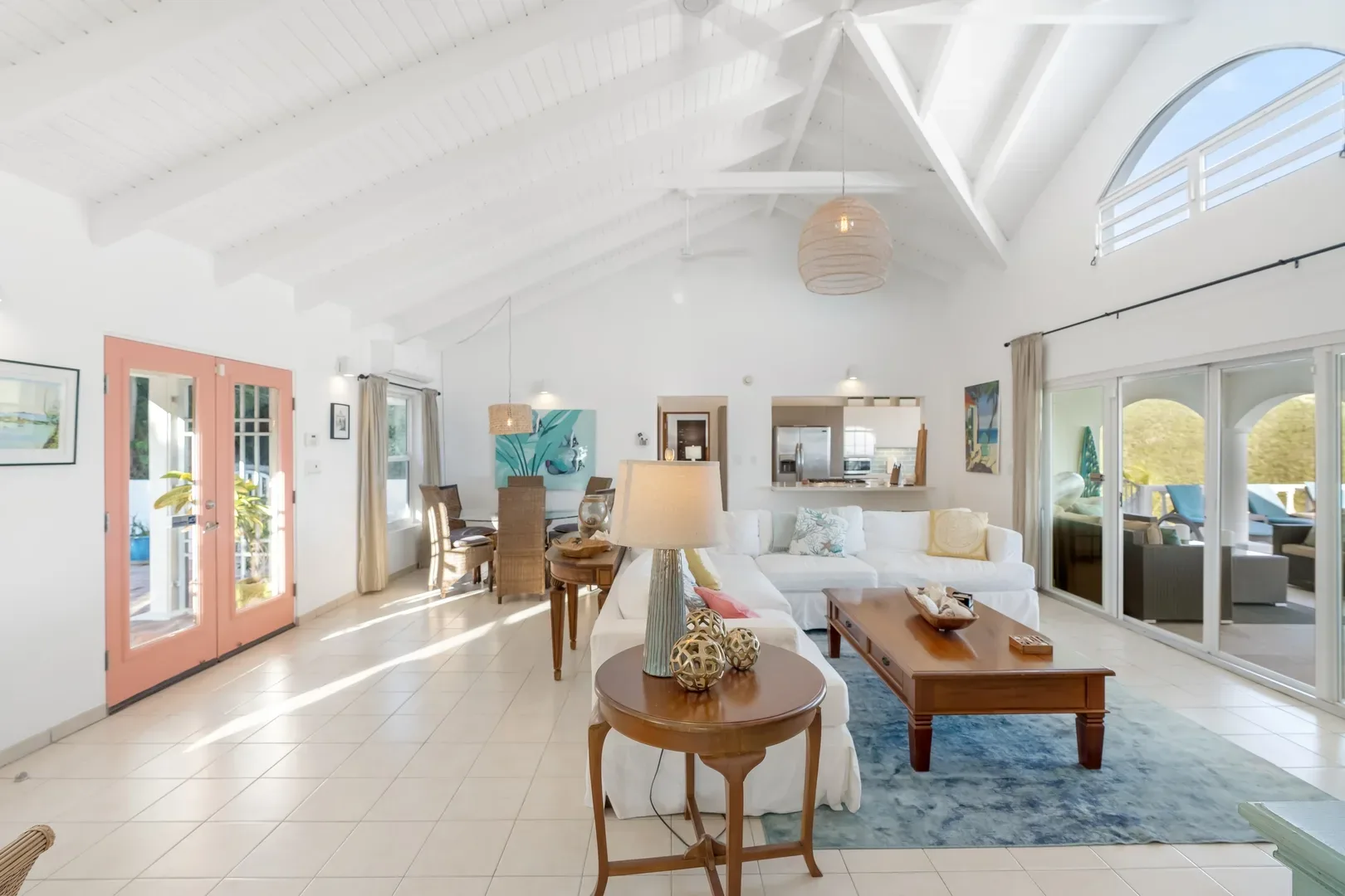 Spacious open-concept living room in St. Maarten villa with white vaulted ceiling, coral French doors, white sofas, wooden furniture, and sliding glass doors to outdoor patio