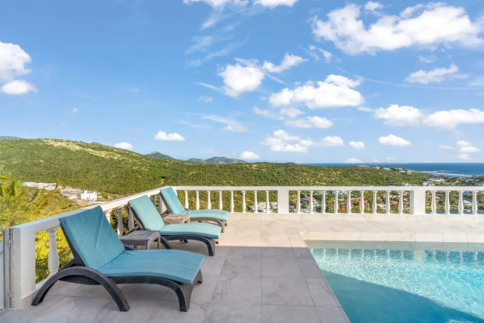 Terrace with turquoise lounge chairs, pool, and panoramic view of green hills and ocean in St. Maarten