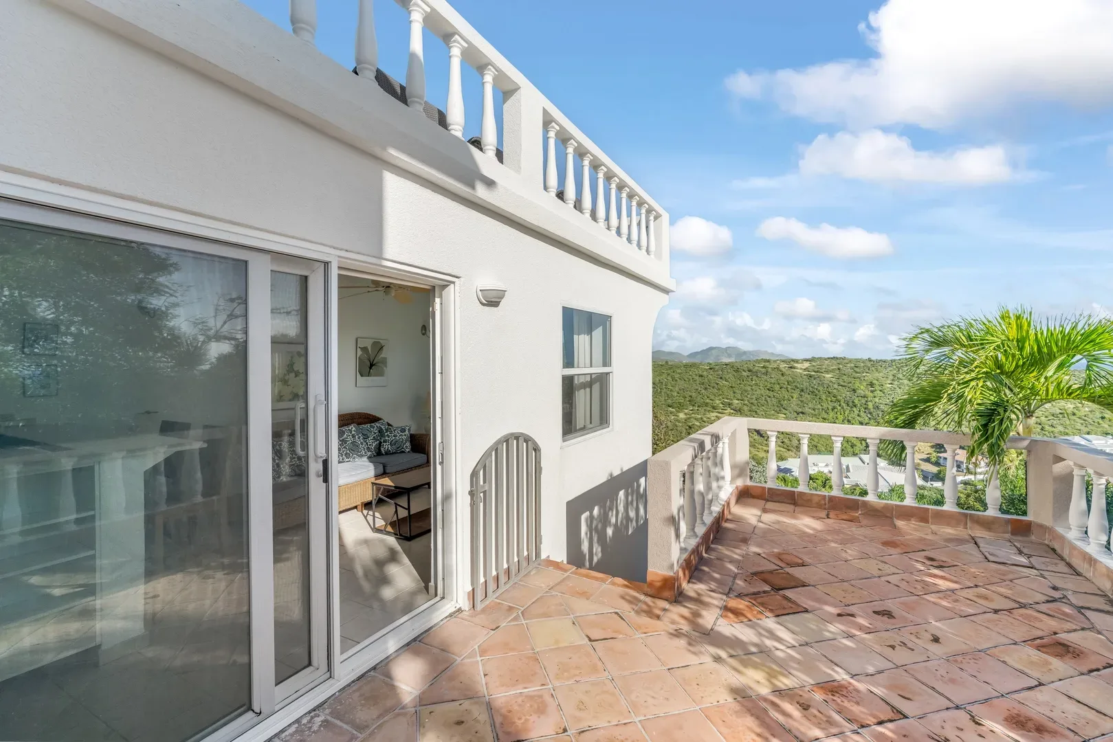 White villa with terracotta-tiled terrace, sliding glass doors leading to a living area with wicker sofa, mountain and green hill views, palm tree, and blue sky