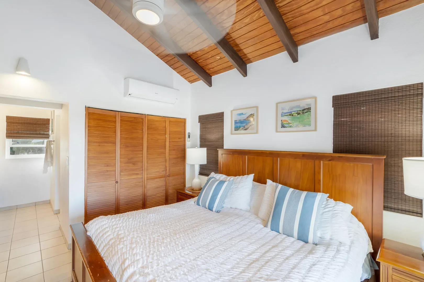 Bright bedroom with a king bed, white quilt, blue-striped pillows, wooden ceiling beams, bamboo blinds, and a view into an adjacent tiled room