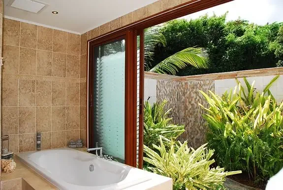 The indoor bathtub sits beside a private doorway opening to a lushly landscaped outdoor shower area for a tropical bathing experience.
