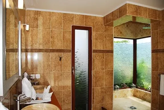 The tan tiled bathroom offers double stone vessel sinks, a frosted glass door, and walk-in shower with large windows overlooking lush greenery.