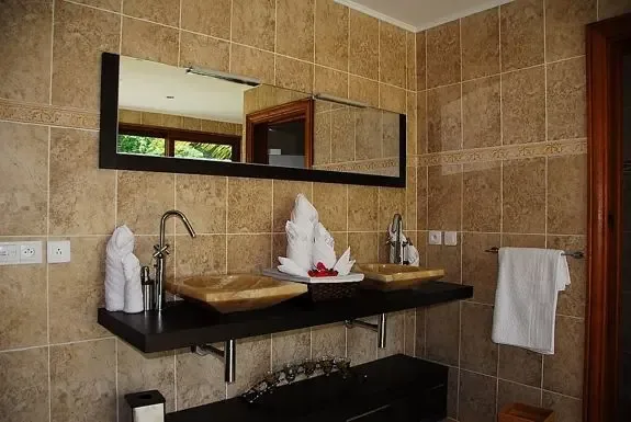The modern double vanity features stone vessel sinks, tall chrome faucets, and decorative towel sculptures against warm tan wall tiles with a decorative border.