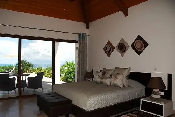 Wake up in this modern bedroom with high vaulted wood ceiling, large bed with decorative pillows, and large glass doorways leading to a terrace with breathtaking ocean views.