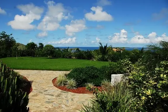 A stone-paved path leads to a manicured green lawn bordered by lush tropical landscaping, offering stunning panoramic views of the blue ocean and coastline.