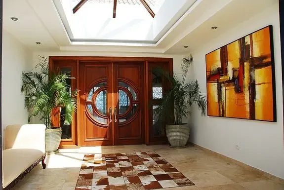 The grand entrance showcases ornate circular-patterned wooden doors, flanked by potted palms, abstract wall art, a checkered rug, and bright natural light from the skylight.