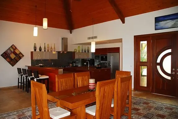 An open-plan kitchen and dining space features a large wooden dining table, breakfast bar with stools, high vaulted wood ceilings, contemporary pendant lighting, and a unique designer front door with circular glass inlay.