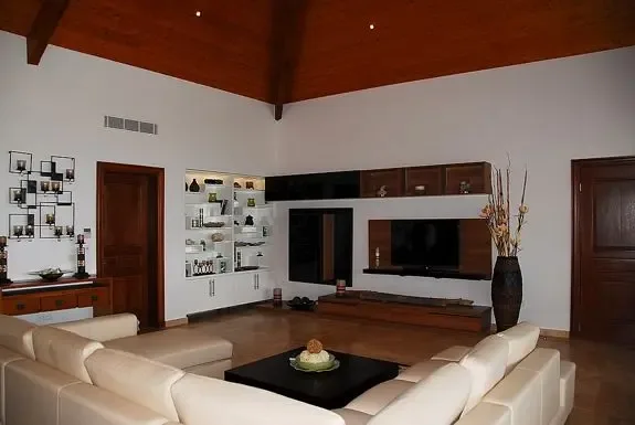A high-ceiling living area features a white L-shaped sectional, modern entertainment unit with built-in TV, wood paneling, and a large decorative vase.