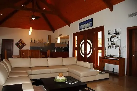 The spacious living room showcases a cream-colored sectional sofa, dark wood coffee table, and high vaulted wood-beam ceilings opening to the dining and kitchen areas with distinctive circular-patterned wooden doors.