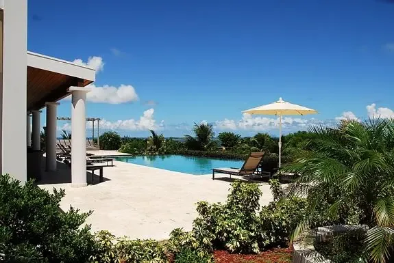 Your private infinity pool area features a stone patio, lounge chairs, and parasol framed by white columns and lush tropical greenery with stunning ocean views.