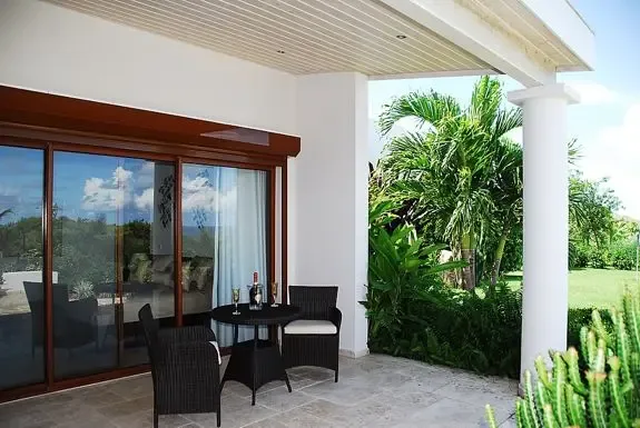 A covered outdoor patio features wicker seating and a small dining table with views of lush tropical landscaping through large glass doorways.